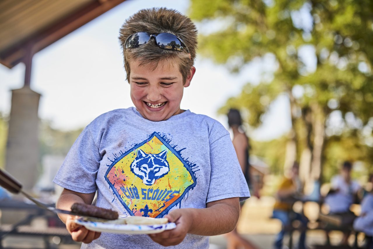 A scout grabs a sausage off the grill.